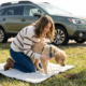 A woman kneels on a travel mat in a grassy roadside area, guiding a new puppy on a harness during a safe potty break stop with a green SUV parked in the background.
